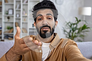 Close-up photo of a smiling young Indian man at home and talking on a video call on the phone to the camera, gesturing