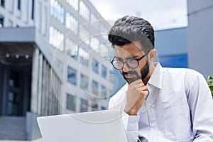 Close-up photo of a thoughtful and serious young Indian businessman in a white shirt sitting outside and staring blankly