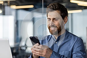 Close-up photo of a tired young man sitting at his desk in the office and using a mobile phone