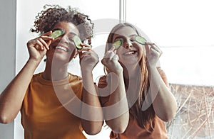 Close up photo of two best girlfriends spending time together having fun while applying cucumber slices on eyes holding them with