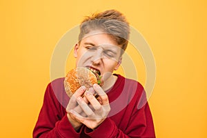 Close-up photo of a very hungry young biting burger with his eyes closed on a yellow background. Hungry student eats quick food,