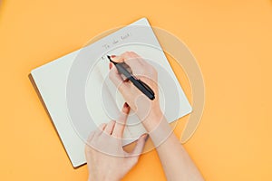 Close-up photo of a woman`s hand writing a to-do list in a clean notebook on an orange background