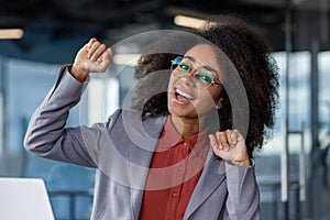 Close-up photo of a young African-American woman in the office, sitting at the workplace and rejoicing in success