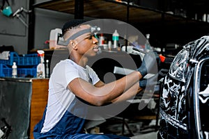Close up photo of young African worker man in protective gloves, cleaning car headlights with foam and special brush
