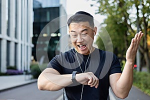 Close-up photo. Young asian male sportsman standing on city street after running and looking at smart watch on hand