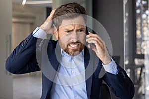 Close-up photo of a young businessman man in a suit standing in an office center, worriedly talking on the phone and