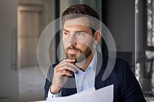 Close-up photo of a young businessman sitting in a modern office in a suit, holding papers in his hands and seriously