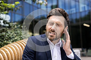 Close-up photo of a young businessman writhing in pain, sitting on a bench near the office and holding his hand by his