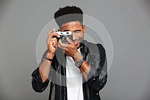 Close-up photo of young cheerful african man looking through retro cameras objective while taking photo