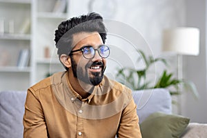 Close-up photo of a young Indian man in glasses sitting on the couch at home and looking to the side with a smile