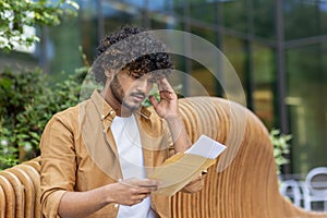 Close-up photo of a young Indian man sitting on a bench outside and sadly reading a letter in an envelope, worriedly