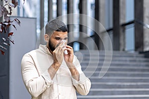 Close-up photo of a young Indian man standing near a building on the street and wiping his nose with a tissue, suffering