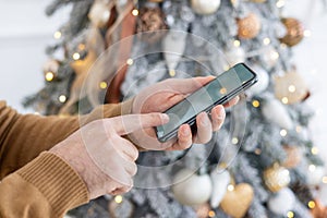 Close-up photo of young male hands holding and using a mobile phone on New Year's holidays near the Christmas tree