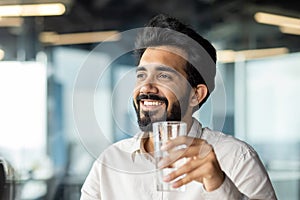 Close-up photo of a young man, an Indian businessman sitting in the office and holding a glass of clean water in his