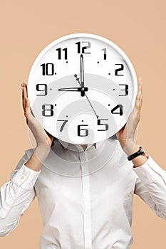 Close-up photo of young modern business woman hold a clock in front of the face over beige background.