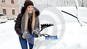 Close up photo of young preaty smiling blond woman in brown coat and jeans trying to clean up snow covered car by blue