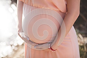 Close up photo of young pregnant woman in elegant pink dress touching her belly with hands.