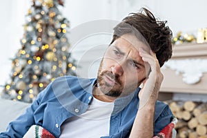 Close-up photo of a young tired man sitting at home on a holiday and not feeling well. He holds his head with his hand