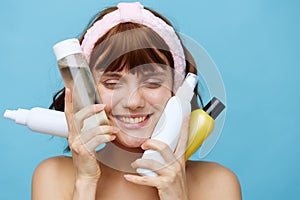 a close-up photo of a young woman with a soft pink headband on her head, holding many different jars of care cosmetics