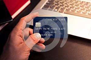 Close up photography of hand`s man holding a global blue credit card with the laptop behind on a wood table. Sunlight photography