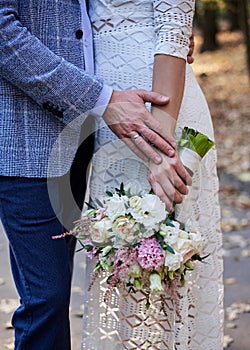 Close-up picture of bride`s and groom`s hands, holding each other and beautiful white and pink flower rose bouquet. Close-up