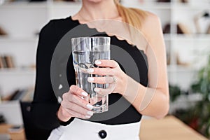 Close-up picture of huge glass of water in hands of young blond woman, standing in office room. Manager at workplace. Work process