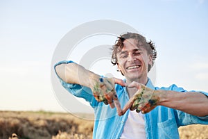 Close-up picture of male artist showing heart sign with dirty with paint hands on wheat field in summer.Painting workshop in rural