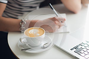 Close up picture of woman`s hands and a cup of cappuccino. Lady is writing in her notebook with a laptop nearby.