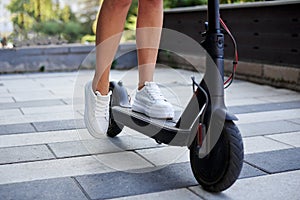 Close-up picture of woman`s legs, riding on electric scooter in city center. Female, wearing white sneakers. Summer leisure