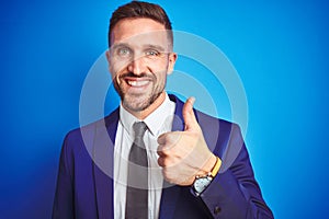 Close up picture of young handsome business man over blue isolated background doing happy thumbs up gesture with hand