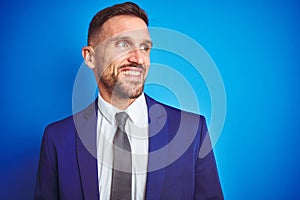Close up picture of young handsome business man over blue isolated background looking away to side with smile on face, natural