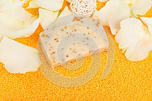 Close-up, a piece of handmade soap on scattered orange bath salts in the form of small balls, surrounded by white rose petals