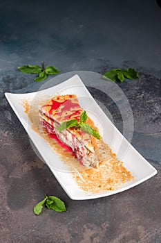 Close-up piece of velvet red cake with cream on white plate decorated with strawberry jam and mint on gray background