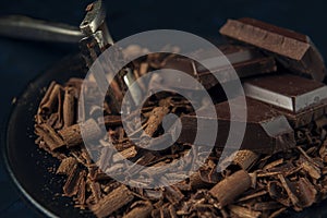 Close-up pieces of chocolate, chocolate shavings and peeler in black plate on dark textured background