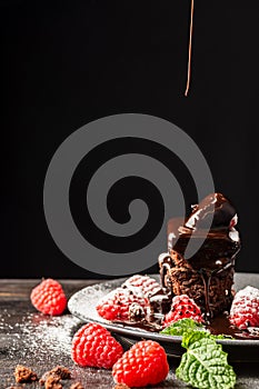 Close-up of plate with brownie, raspberries and mint leaves with falling chocolate syrup, on wooden table, selective focus, black