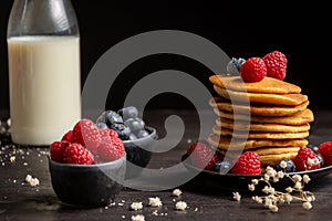 Close-up of plate with stacked pancakes, blueberries, raspberries and bottle of milk, on table and dark background