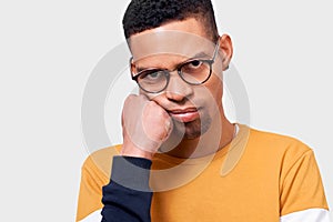 Close-up poratrait of tired Afro male leans on hand, has displeased expression, wearing sweaterand eyewear, posing in studio.