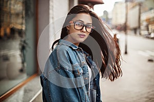 Close-up portrait of adorable girl posing on the street. girl student in a denim jacket and glasses looking in camera