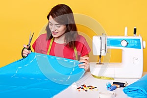 Close up portrait of adorable seamstress wears red t shir, sitting at table. Dressmaker cuts dress detail on the sketch lines,