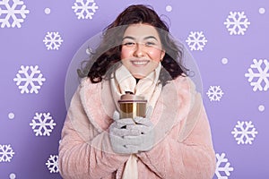 Close up portrait of adorable smiling woman wearing pale pink faux fur coat and, holding thermo mug with coffee or tea, drinking