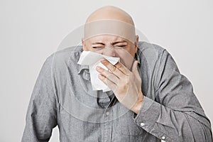 Close-up portrait of adult bald caucasian man sneezing in napkin and shrugging, standing with closed eyes over gray