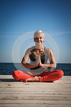 Close-up portrait of adult blond woman with short haircut practices yoga on the pier against the background of the sea and blue