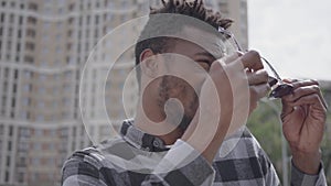 Close up portrait of African American man standing against the background of urban building squinting in the sun, then