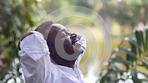 Close up portrait of african business man with calm face close eyes stand on green nature background with hands above head