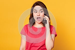 Close up portrait of amazed beautiful girl in casual style, has dark hair, standing and speaking on mobile smart phone and looking