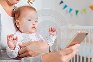 Close up portrait of a amazement baby sitting in mother`s arms and looking at the smartphone that she uses