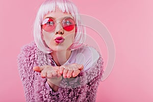 Close-up portrait of amazing female model in pink wig sending air kiss. Studio photo of good-humour