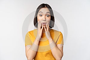 Close up portrait of asian woman looking surprised, wow face, staring impressed at camera, standing over white