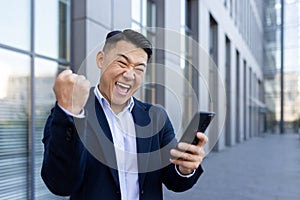 Close-up portrait of an Asian young business man standing in a suit near an office building, holding a phone in his