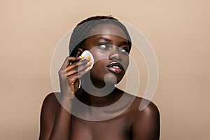 Close up portrait of an attractive african american woman removing makeup with sponge isolated on beige background